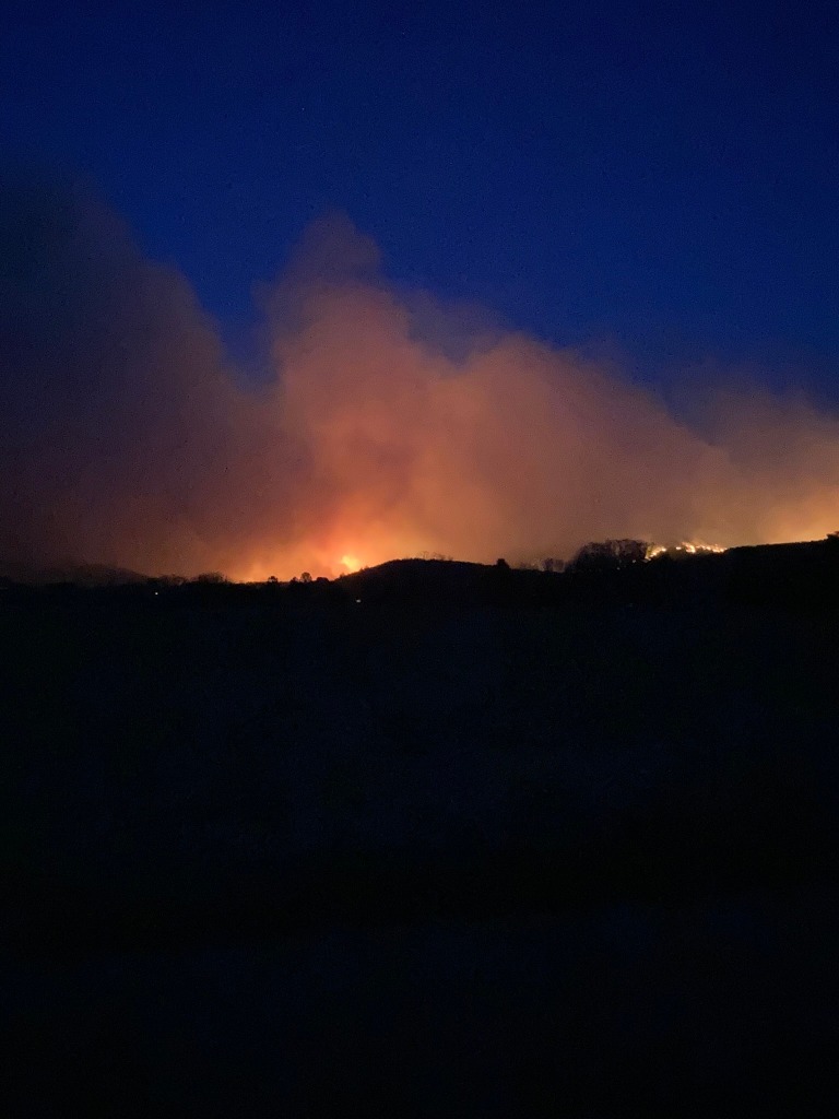 A photo taken just after sunset--a ridge line appears silhouetted in black against a deep blue sky. Bright orange and yellow flames are visible along the top of the ridge, and there is much smoke coming off the fires. The smoke is lit up by the fires, purple and orange.